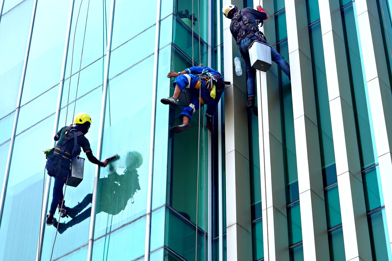 home-hero-bg Three window cleaners suspended on ropes clean a modern glass building facade during the day.
